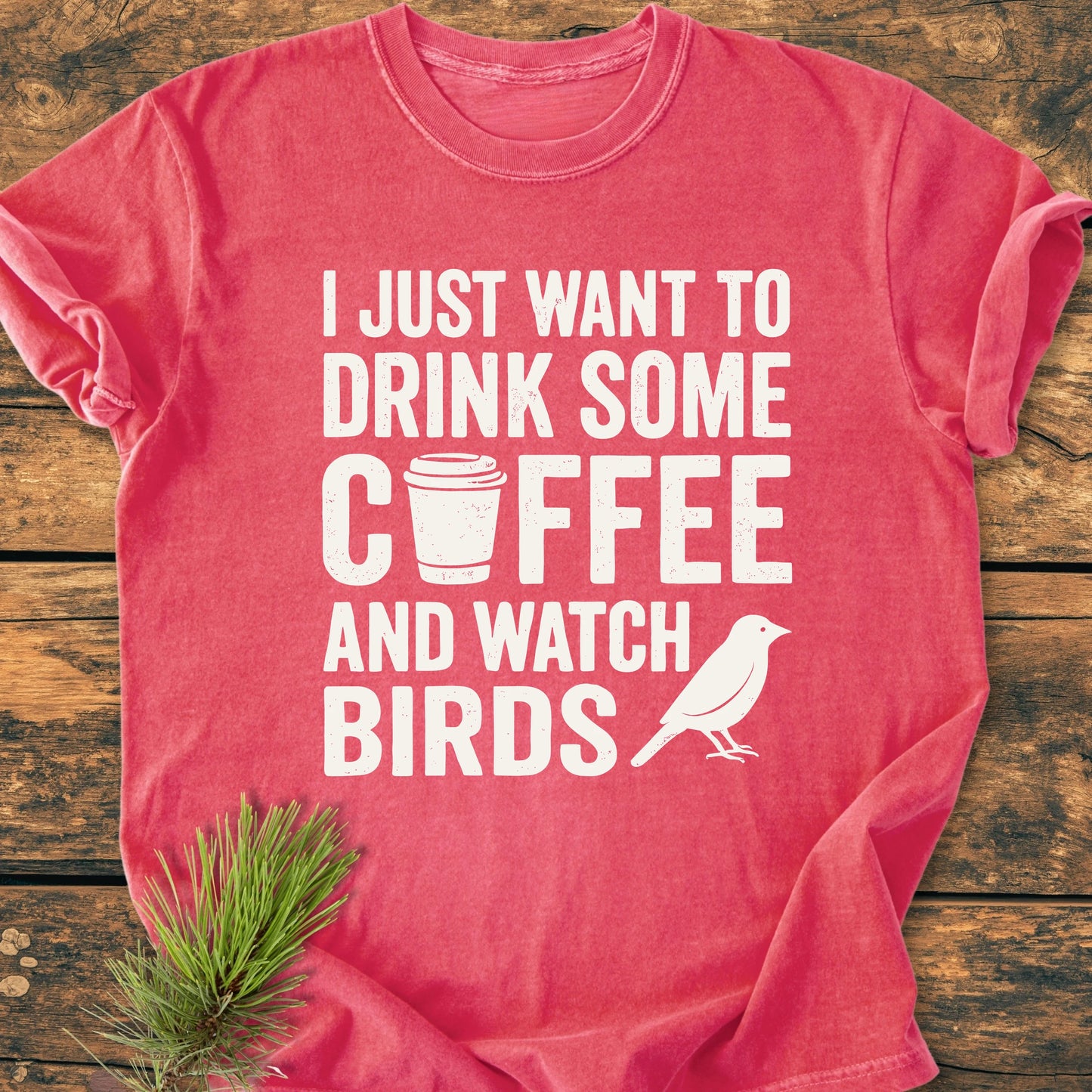 Coral-red 1405 Drink Coffee And Watch Birds Base T-shirt with coffee cup and bird graphics, laid on wood. Features bold white text, pine needle sprig, and showcases Comfort Colors' soft-washed, relaxed fit design.