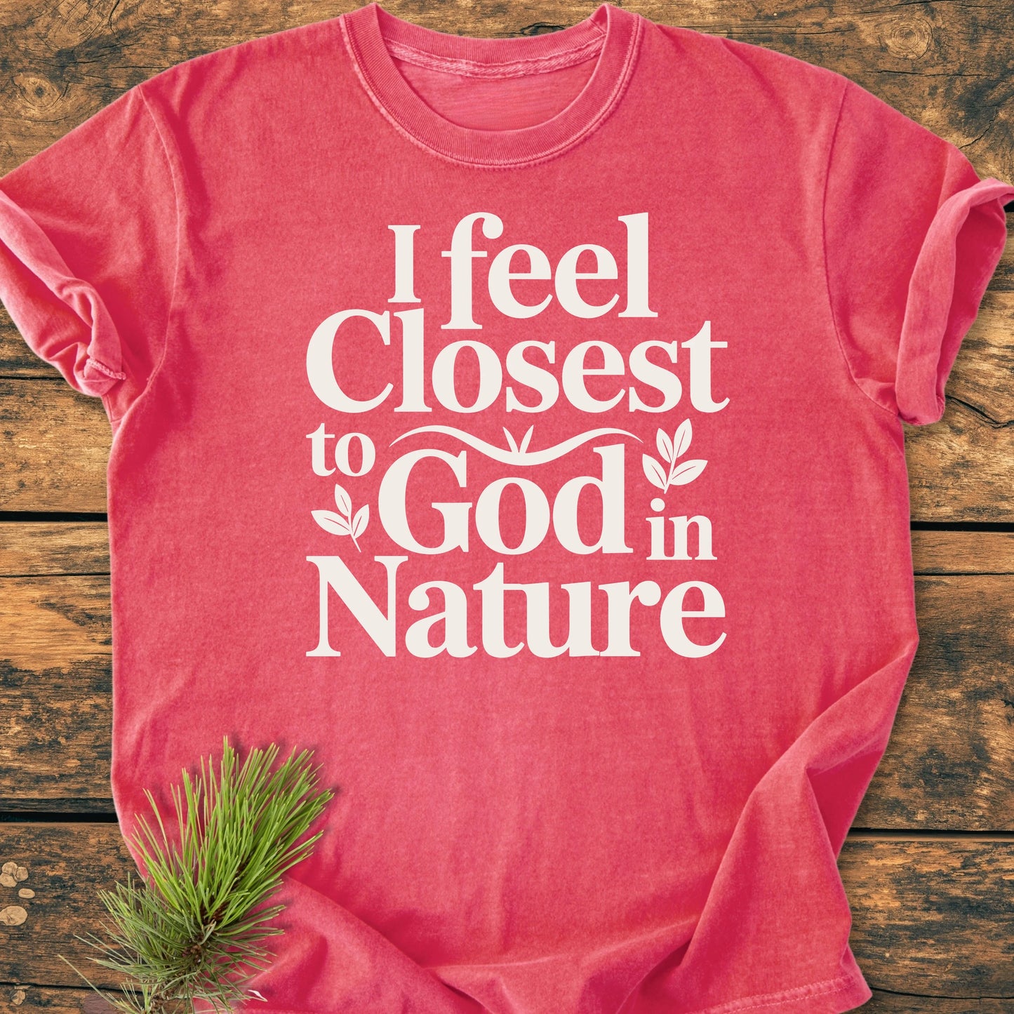 1427 Closest To God - Vintage Wash Tee: Pink-red T-shirt with white slogan and leaf graphics, laid flat on wood, featuring a small pine sprig. Designed for a relaxed, lived-in feel.