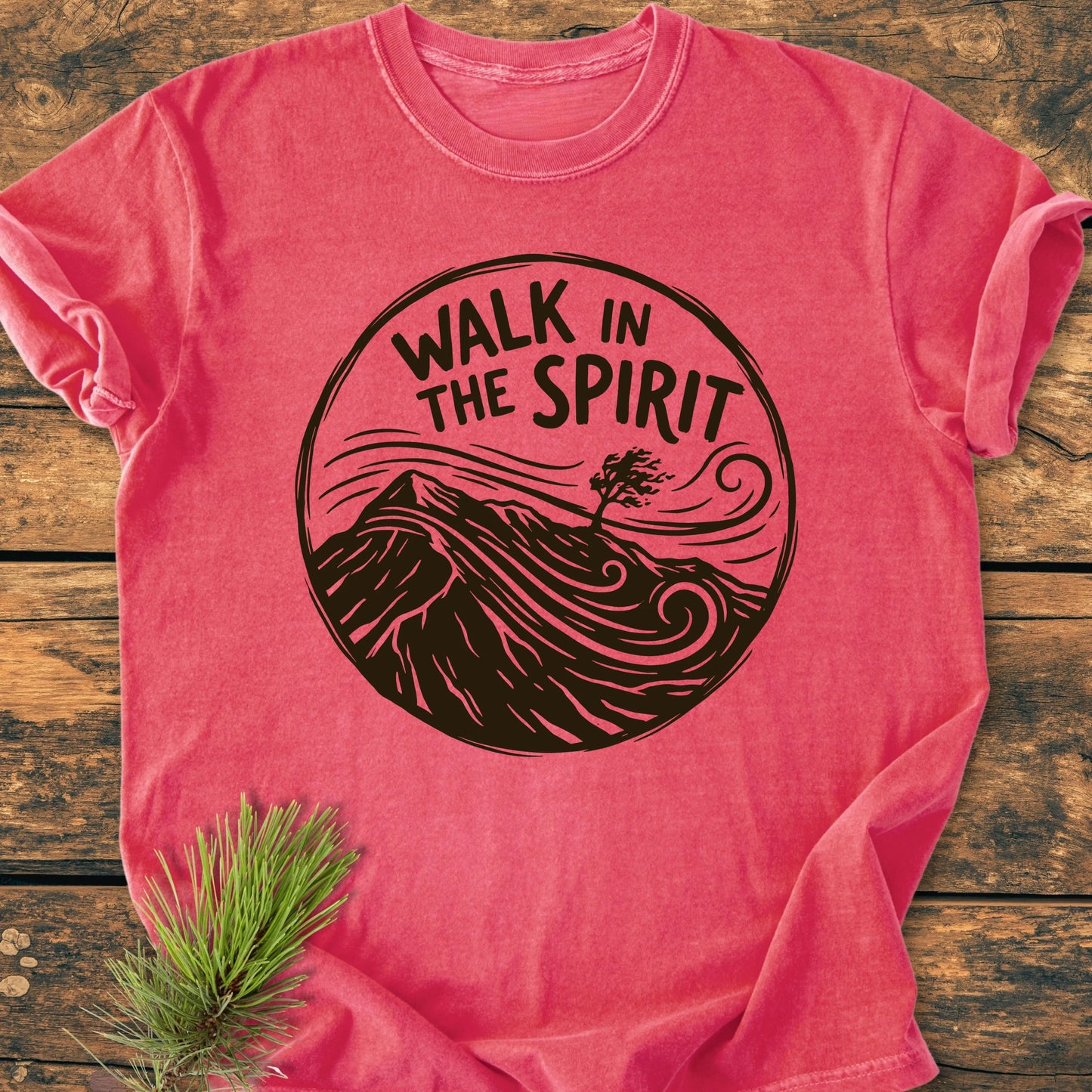 1410 Walk In The Spirit T-shirt features a circular graphic with hills, tree, and wind lines, printed on a soft, relaxed-fit red cotton tee, laid on a wooden surface with pine sprig.