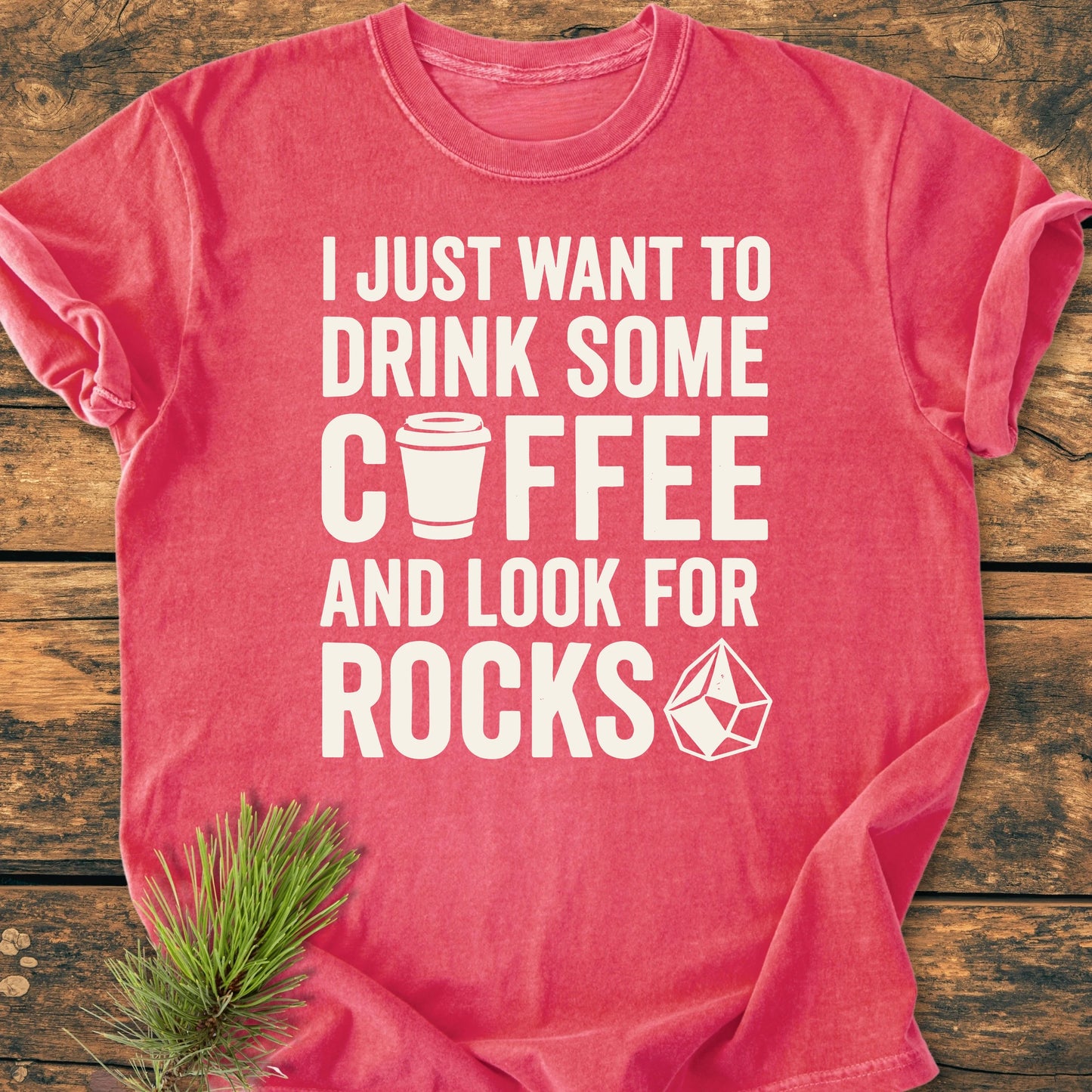 Red-pink 1404 Drink Coffee And Look For Rocks Base t-shirt with white text and icons, laid flat on wood, beside a pine sprig. Comfortable, durable, relaxed fit, 100% ring-spun cotton.