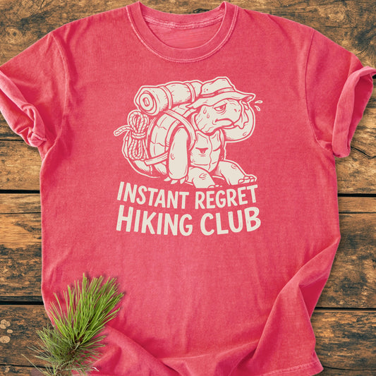 Heathered red T-shirt featuring a cartoon turtle with hiking gear, captioned “INSTANT REGRET HIKING CLUB.” Laid flat on wood with pine sprig. Vintage wash, relaxed fit, durable stitching.