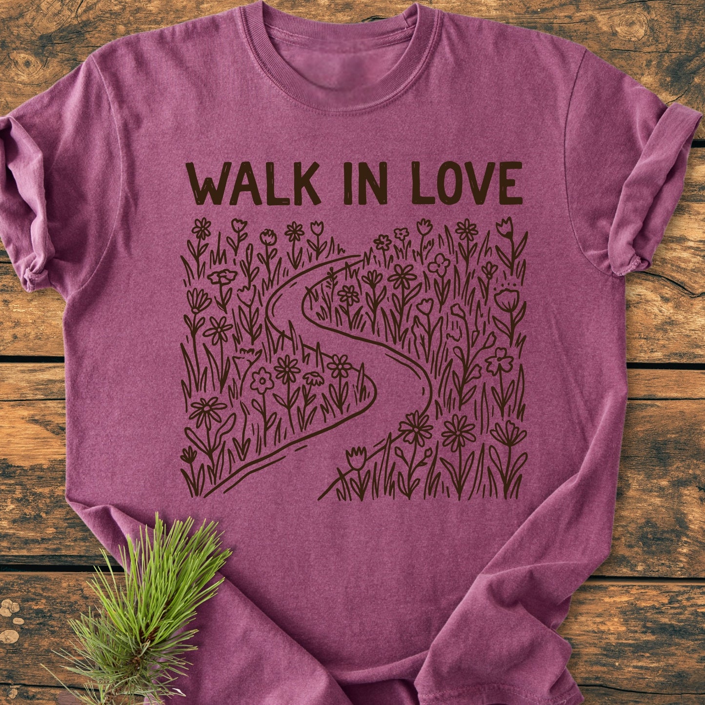 Walk In Love - Vintage Wash Tee features a “WALK IN LOVE” print with a path illustration on a heather-plum background, laid on wood. Includes a sprig of evergreen detail.
