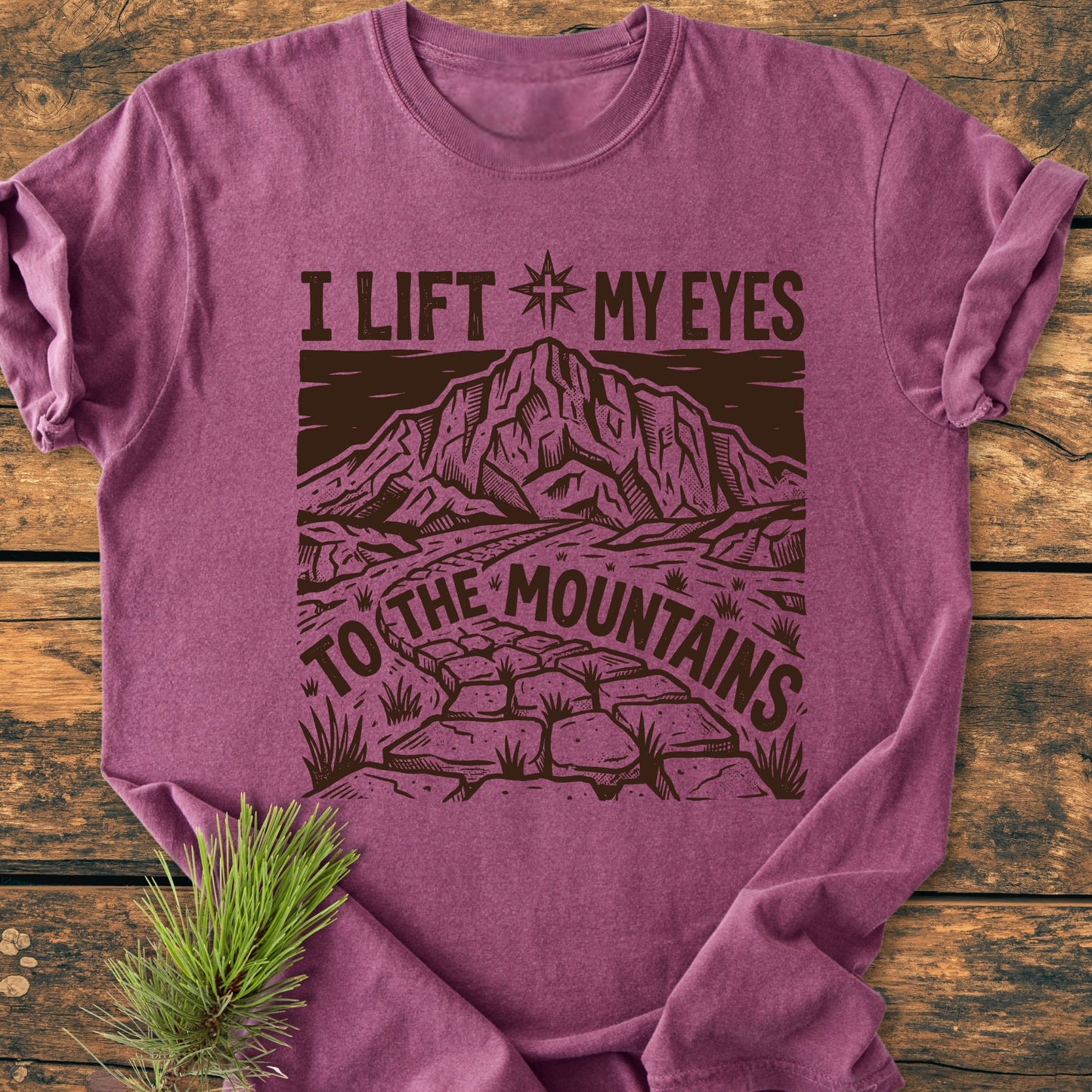 Heather-purple I Lift My Eyes, Psalm 121 - Vintage Wash Tee featuring a mountain scene with a compass and path, laid flat on wood, with a pine sprig beside it.