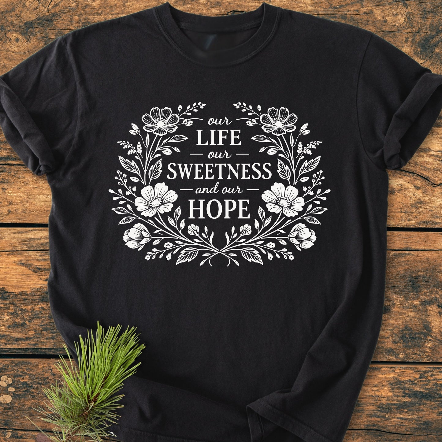 Our Life, Sweetness, Hope - Vintage Wash Tee features a white floral wreath design on a black T-shirt, laid flat on wood with a pine sprig below, highlighting its cozy, relaxed fit.