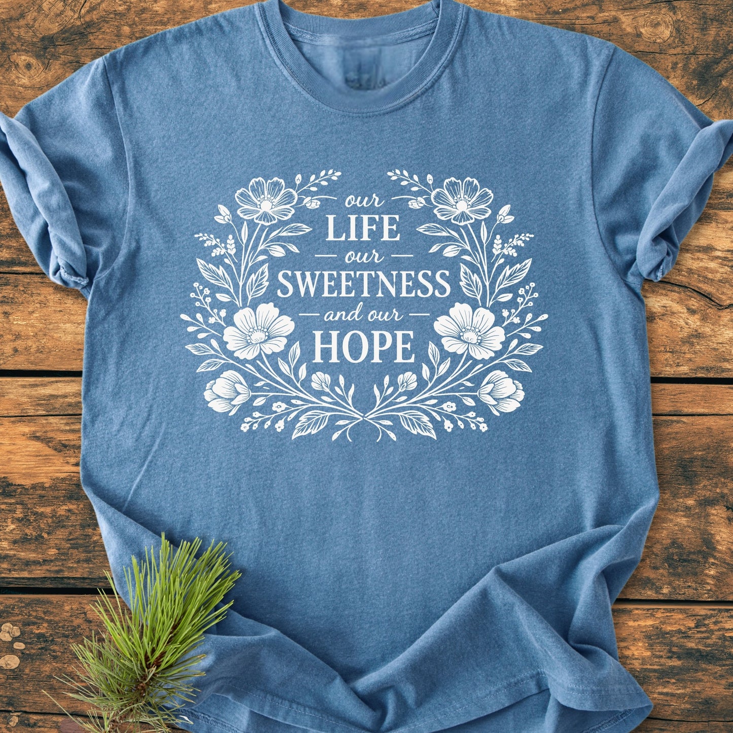 Our Life, Sweetness, Hope - Vintage Wash Tee featuring a white floral wreath design on a heather-blue background, laid flat on wood, highlighting its relaxed fit and soft 100% cotton fabric.