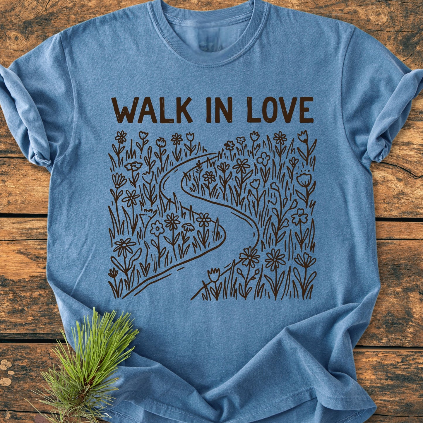 Walk In Love - Vintage Wash Tee featuring bold lettering and a meadow path design, laid on wood with a pine sprig. Made of soft, durable, pre-shrunk 100% ring-spun cotton.