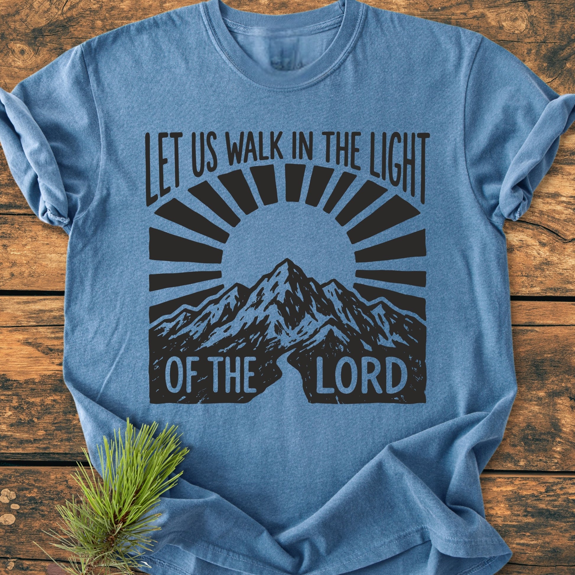 Let Us Walk In The Light Of The Lord - Vintage Wash Tee: Blue heather T-shirt with a sunburst over mountains design, laid on wood with pine sprig. Soft-washed, durable, relaxed fit.