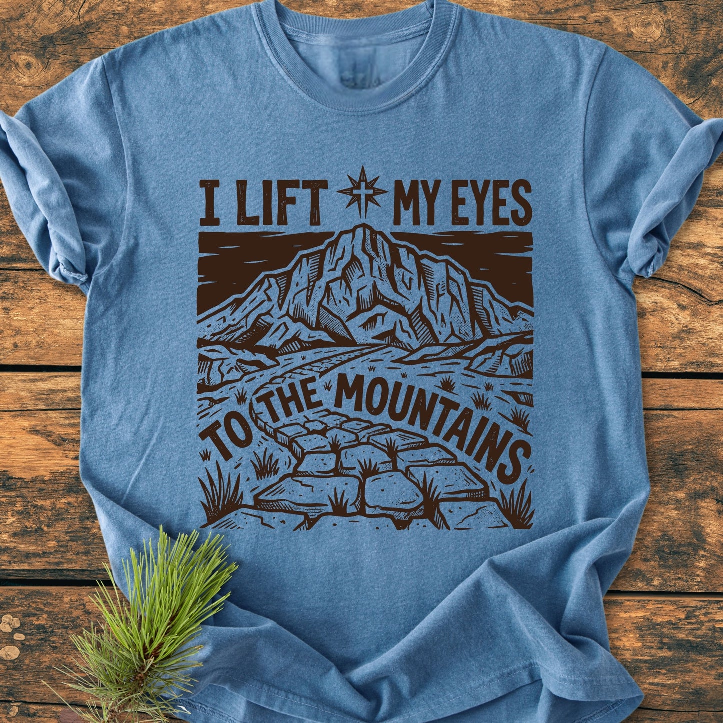 Heather-blue I Lift My Eyes, Psalm 121 - Vintage Wash Tee featuring a mountain scene and compass star, laid flat on wood with a pine sprig; soft, garment-dyed 100% ring-spun cotton.