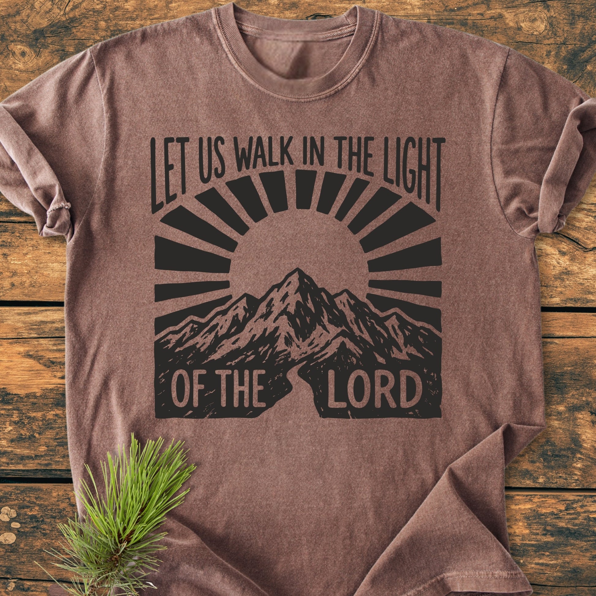 Let Us Walk In The Light Of The Lord - Vintage Wash Tee, folded on wood, features a mountain and sun-rays design. A spruce-pine twig adds an outdoorsy touch, emphasizing its rustic appeal.