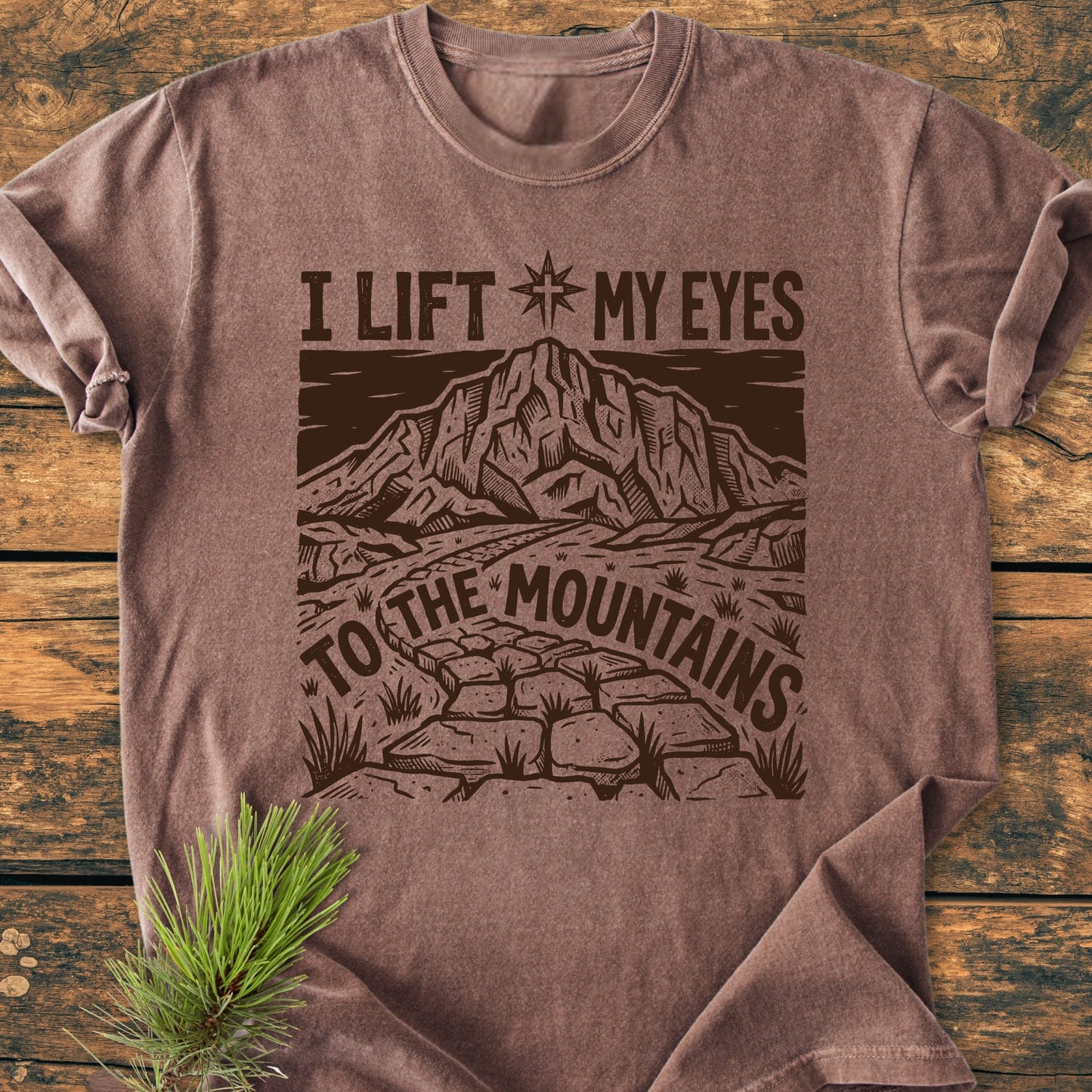 Heather-brown I Lift My Eyes, Psalm 121 - Vintage Wash Tee featuring a mountain scene and text, laid flat on wood with a pine sprig, highlighting its relaxed, durable, and soft-washed garment-dyed cotton design.