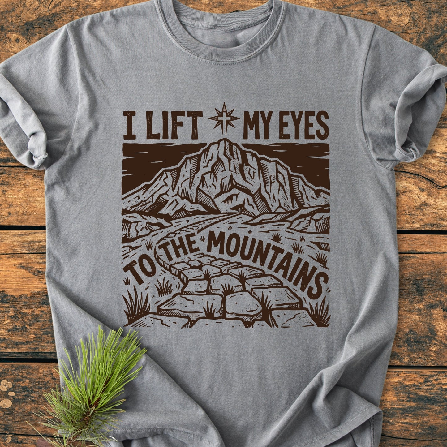 Heather-gray I Lift My Eyes, Psalm 121 - Vintage Wash Tee featuring a stone path, mountain, and star design, styled with a pine sprig on a wooden surface, highlighting its relaxed, durable fit.
