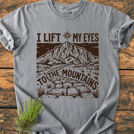 Heather-gray I Lift My Eyes, Psalm 121 - Vintage Wash Tee featuring a stone path, mountain, and star design, styled with a pine sprig on a wooden surface, highlighting its relaxed, durable fit.