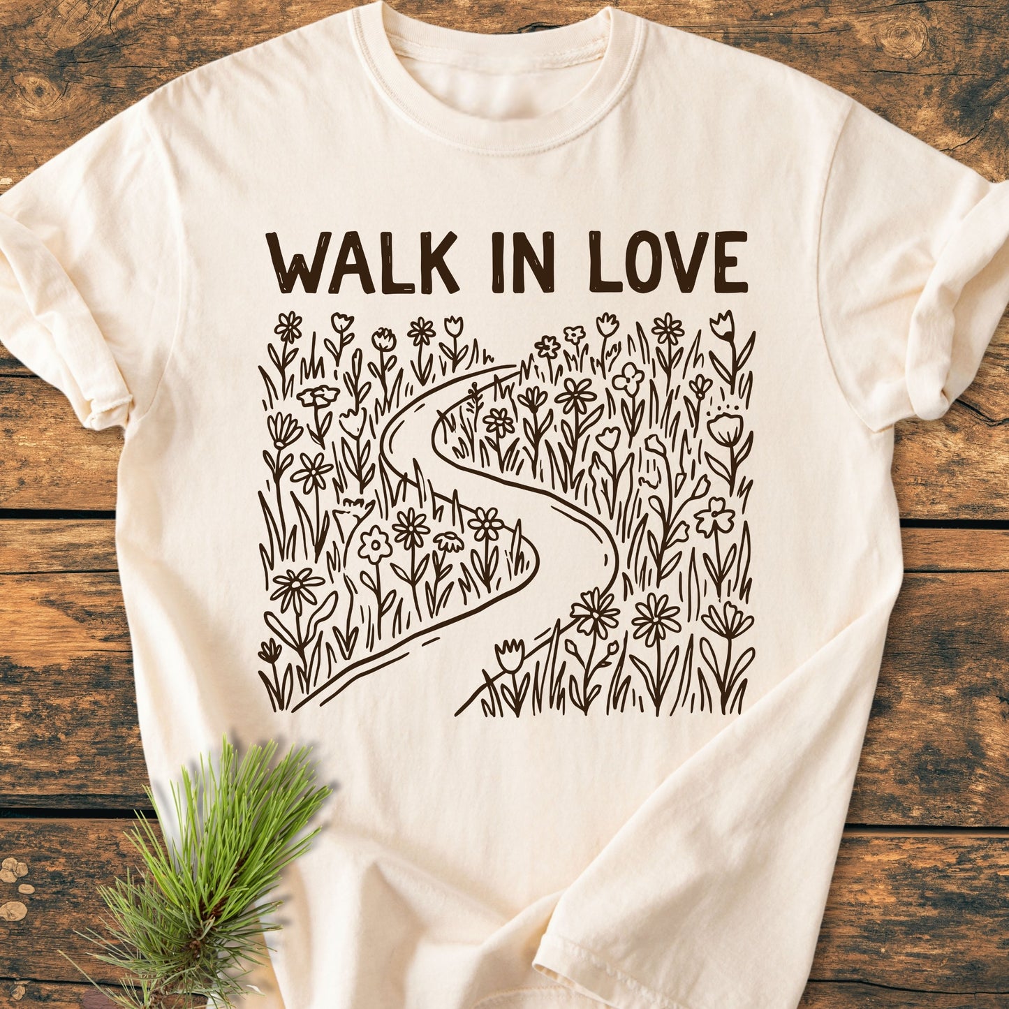 Walk In Love - Vintage Wash Tee features cream hue, bold “WALK IN LOVE” lettering, and line-art of a path with wildflowers, laid on wood with a pine sprig.
