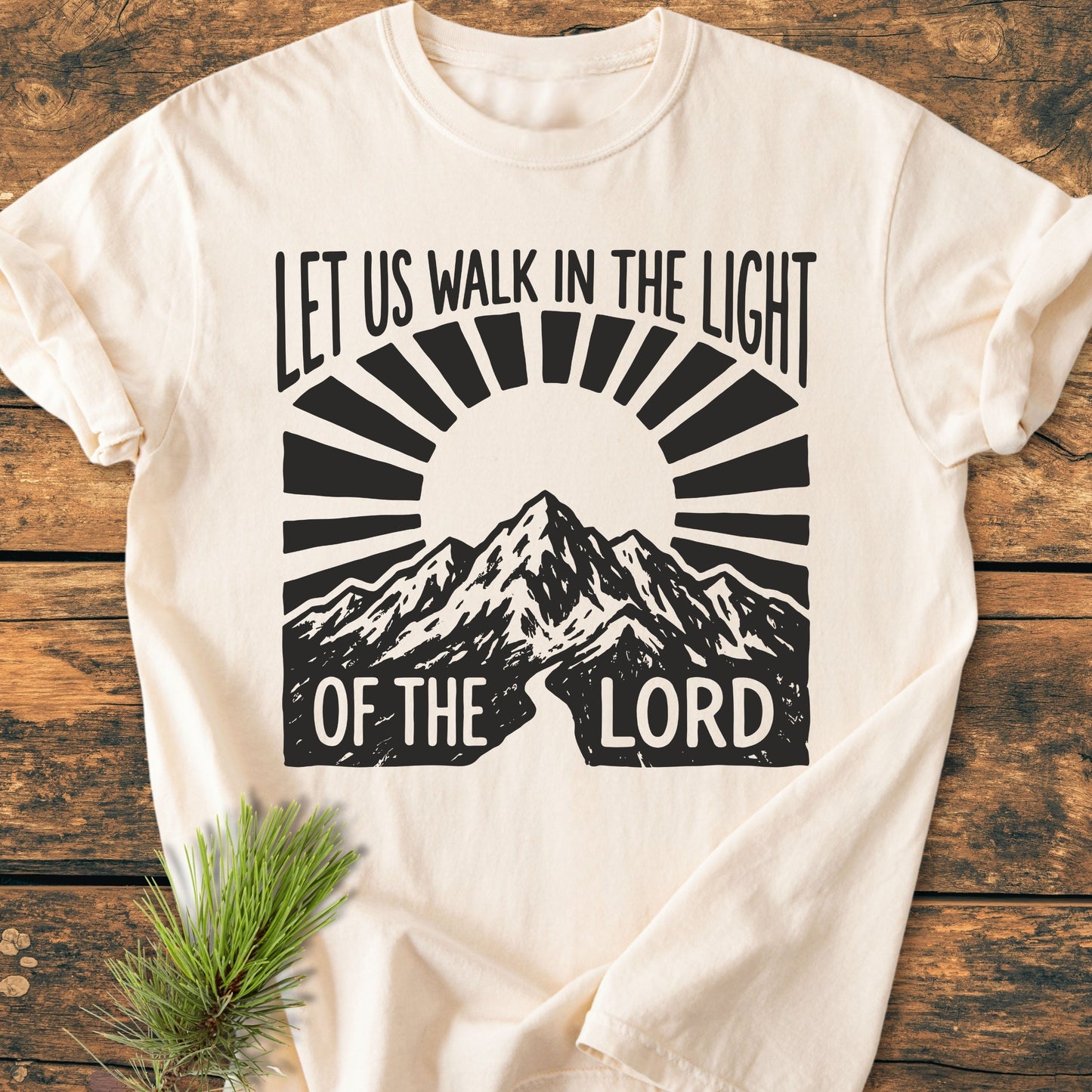 Let Us Walk In The Light Of The Lord - Vintage Wash Tee features a stylized mountain range and sun rays design, laid flat on wood with a pine sprig accentuating its casual, cozy appeal.