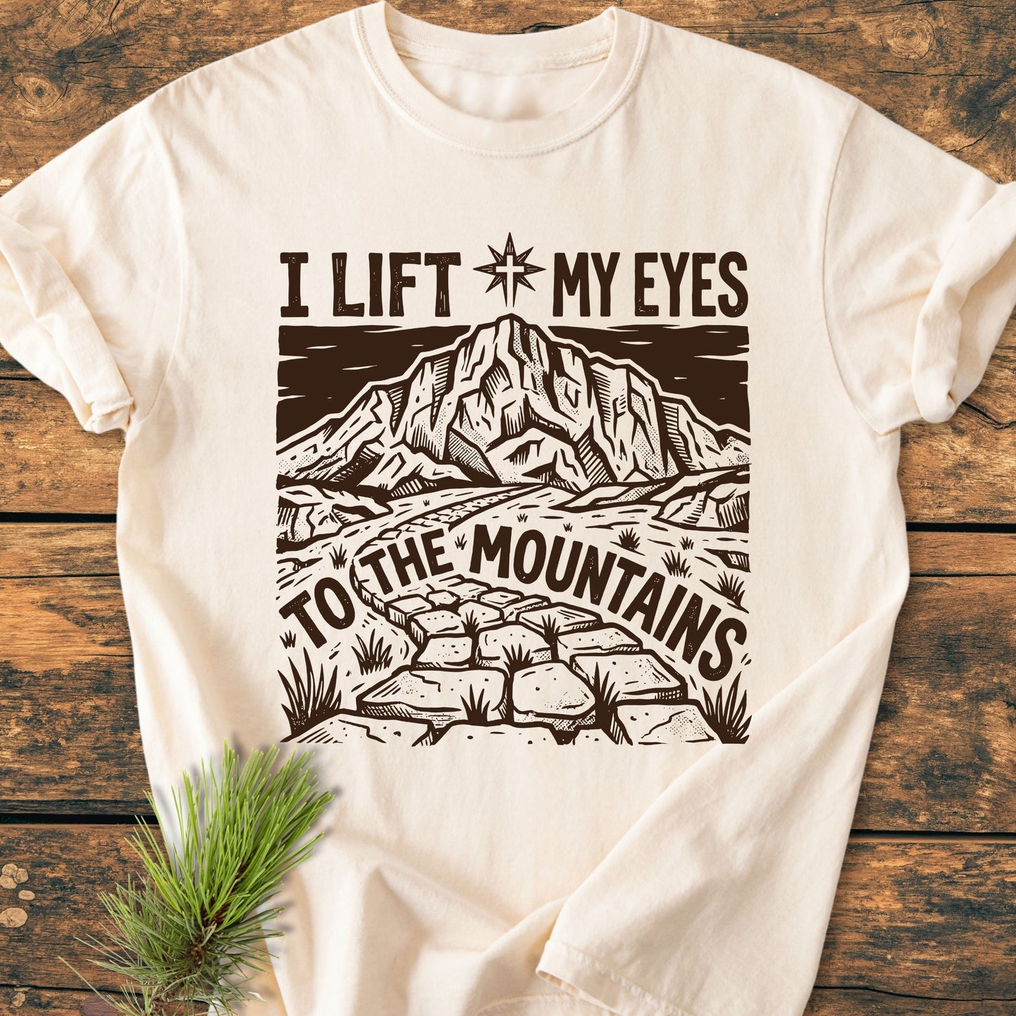 I Lift My Eyes, Psalm 121 - Vintage Wash Tee featuring a stylized mountain and star compass design on light-cream. Laid on wood, pine sprigs accentuate its outdoorsy theme.