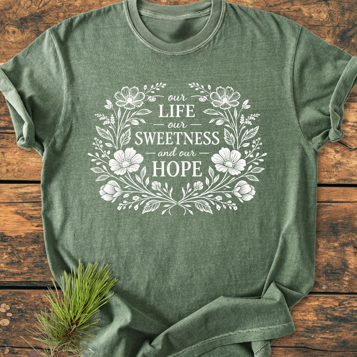 Our Life, Sweetness, Hope - Vintage Wash Tee features a floral wreath encircling inspirational text on a heather forest-green crew-neck, accented by a pine sprig on a wooden surface.