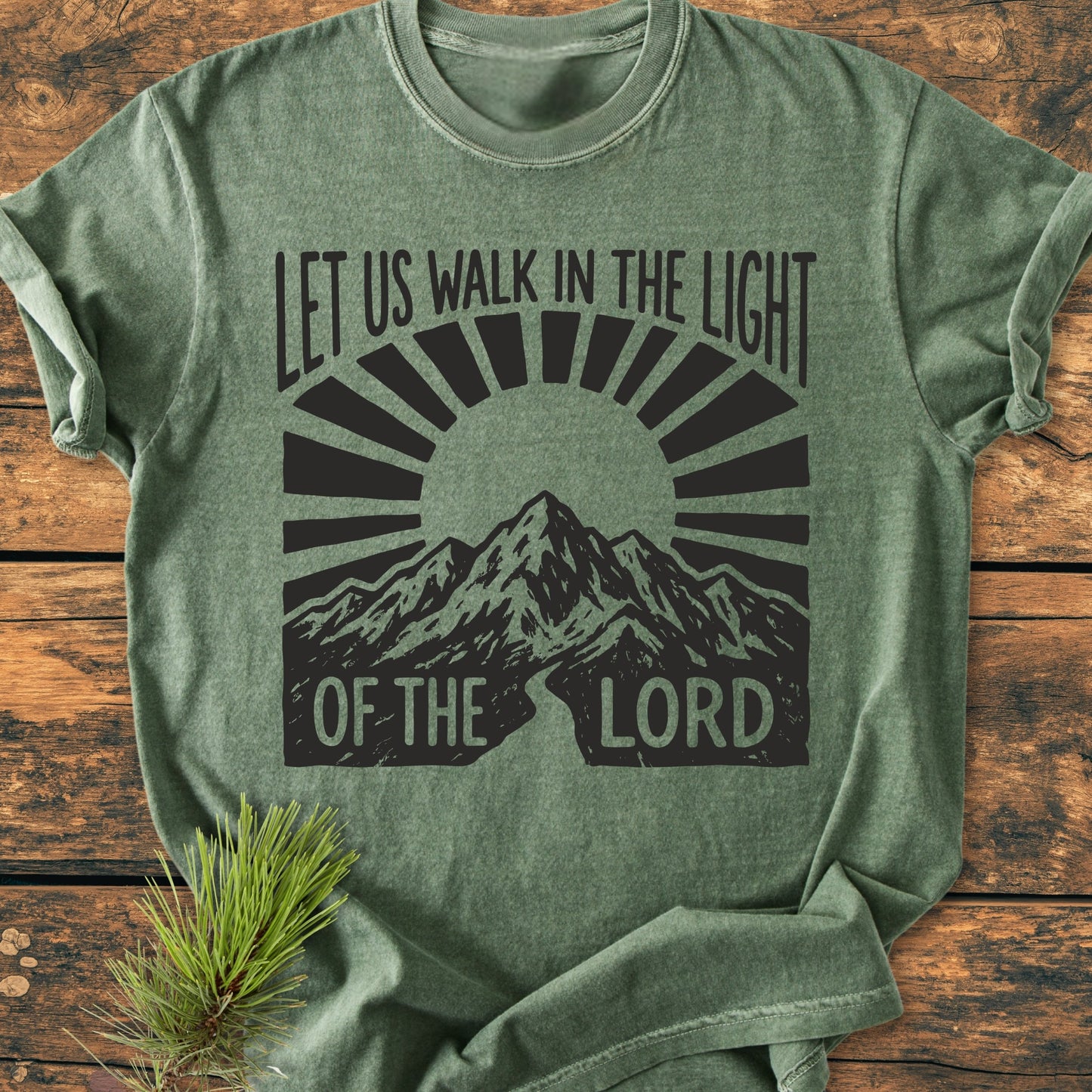 Let Us Walk In The Light Of The Lord - Vintage Wash Tee. Heather-green T-shirt with mountain graphic, sun-rays, and text. Laid on wood with a pine sprig, showcasing durability and comfort.