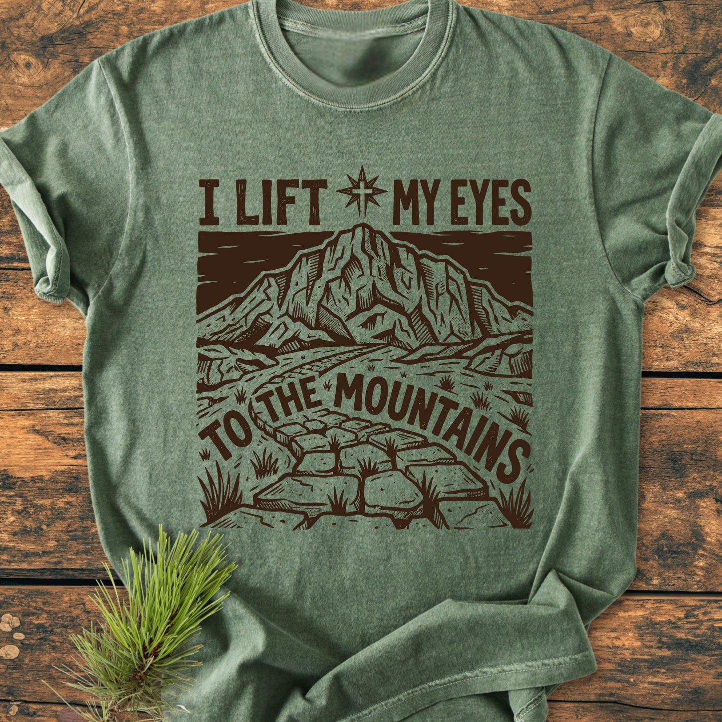 Heather-green I Lift My Eyes, Psalm 121 - Vintage Wash Tee featuring a wood-cut-style mountain graphic and text, laid flat with a pine sprig accent, embodying a rustic, relaxed aesthetic.
