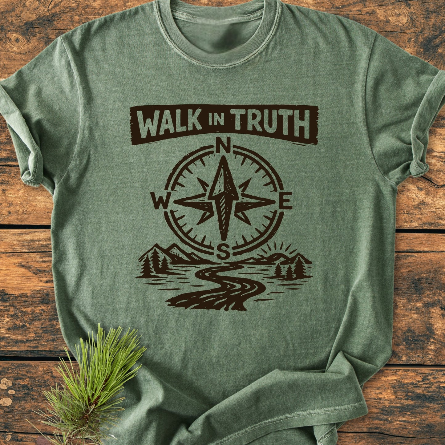 Walk In Truth 3 John 1,4 - Vintage Wash Tee on wooden surface, featuring WALK IN TRUTH text and compass rose over landscape graphic. Pine sprig accentuates the outdoorsy theme.