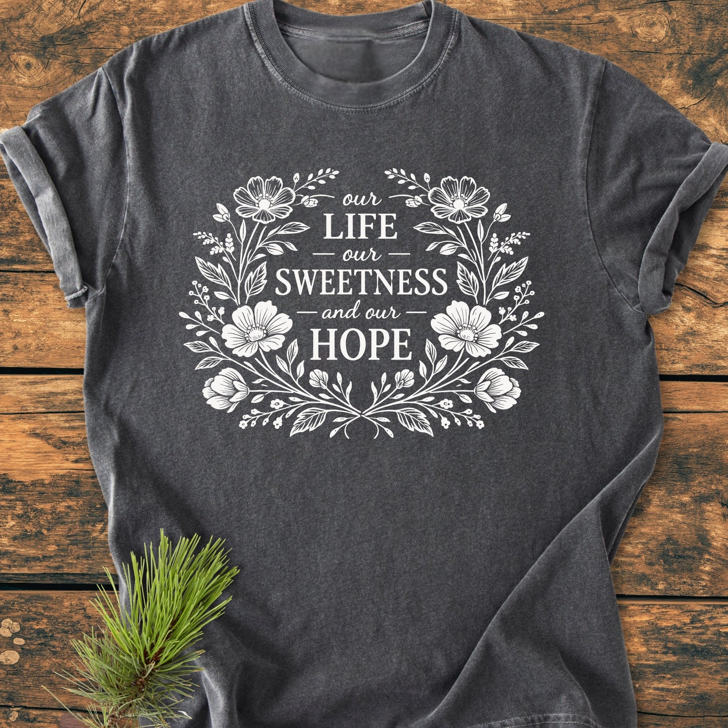 Our Life, Sweetness, Hope - Vintage Wash Tee featuring a white floral wreath design on charcoal-gray fabric, laid on wood with a pine sprig. Relaxed fit, soft-washed, and durable garment-dyed cotton.