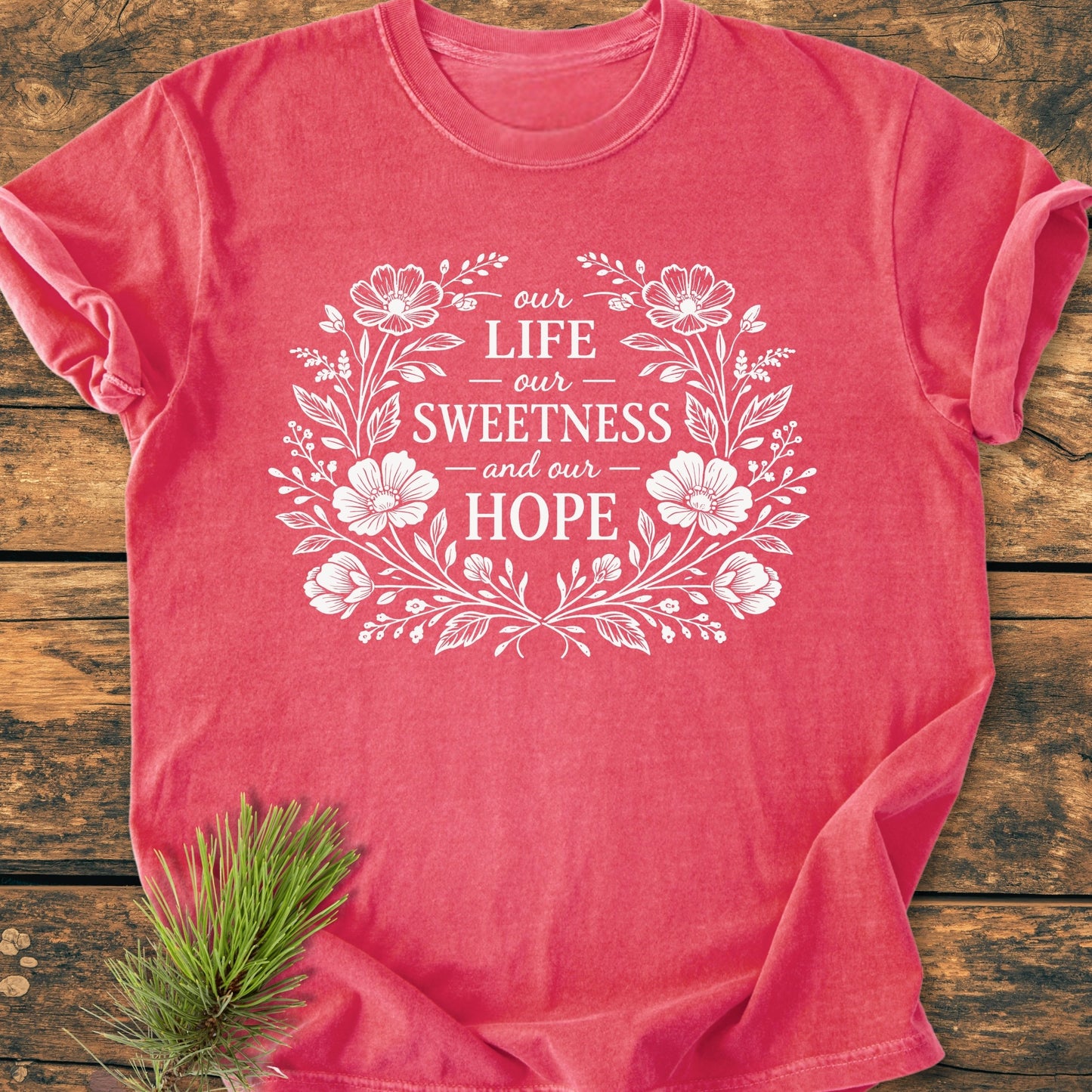 Our Life, Sweetness, Hope - Vintage Wash Tee featuring a floral wreath design on a soft pink/red T-shirt, displayed flat on wood with a pine sprig accenting the corner.