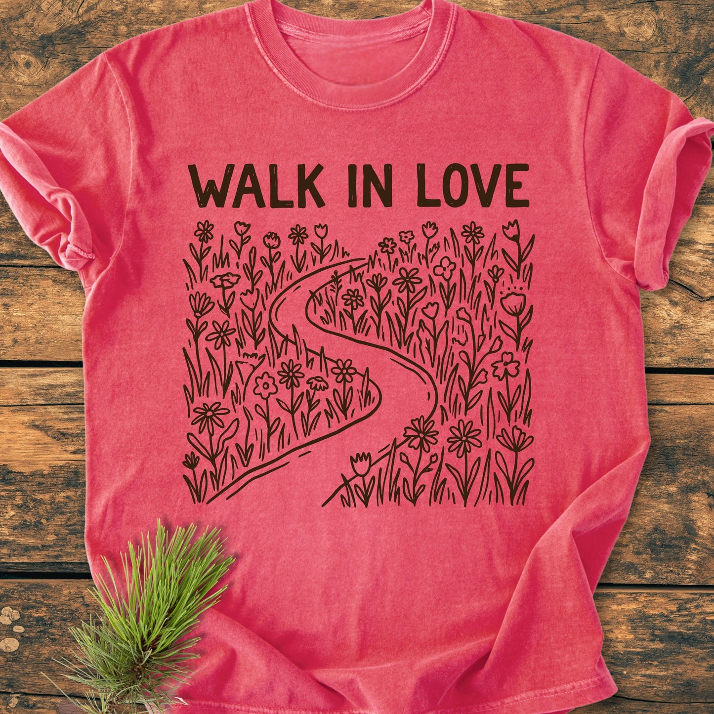 Walk In Love - Vintage Wash Tee features a heather-red design with a black line-drawing of a path through flowers, bold WALK IN LOVE text, laid on a wooden surface with a pine branch accent.
