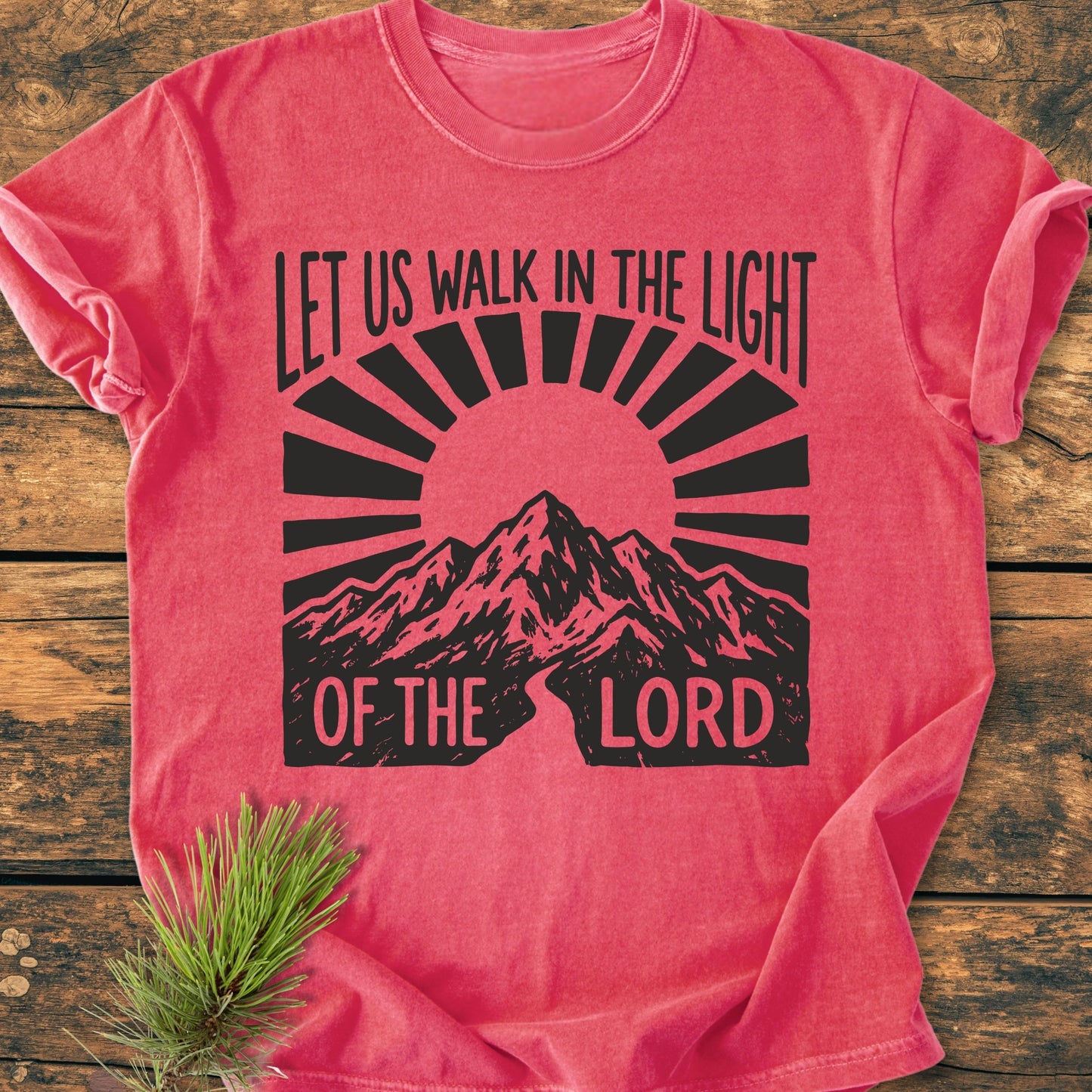 Let Us Walk In The Light Of The Lord - Vintage Wash Tee features a stylized sunburst and mountain design printed on a cozy, relaxed-fit pink-red T-shirt, laid flat with a pine sprig.