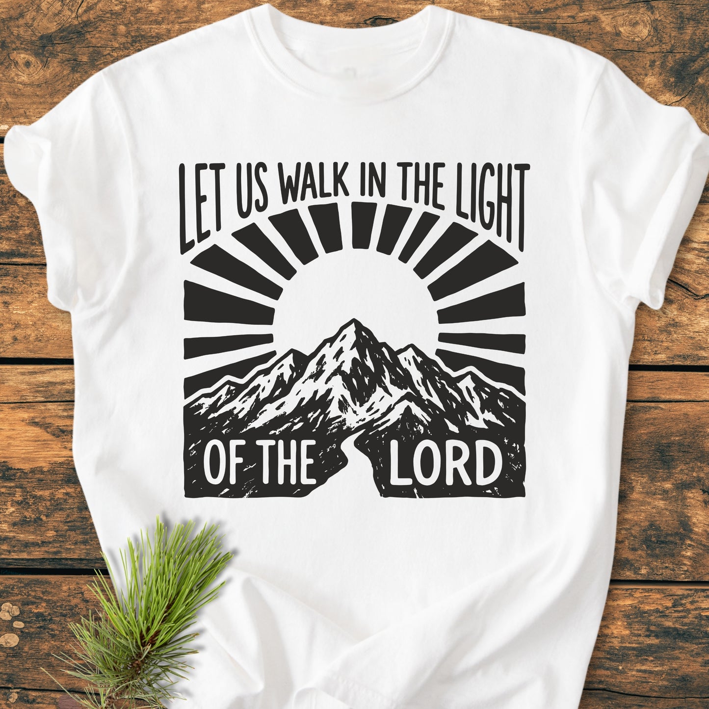 Let Us Walk In The Light Of The Lord - Vintage Wash Tee featuring a mountain range with rising sun graphic, laid flat on wood with pine sprig. Made of soft, durable, 100% ring-spun cotton.
