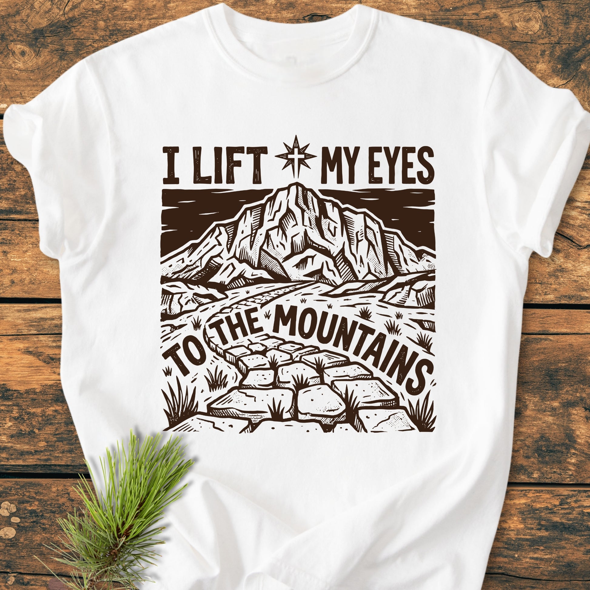 I Lift My Eyes, Psalm 121 - Vintage Wash Tee features a mountain scene with a compass star, laid flat with a pine sprig, highlighting its rustic style and relaxed fit.