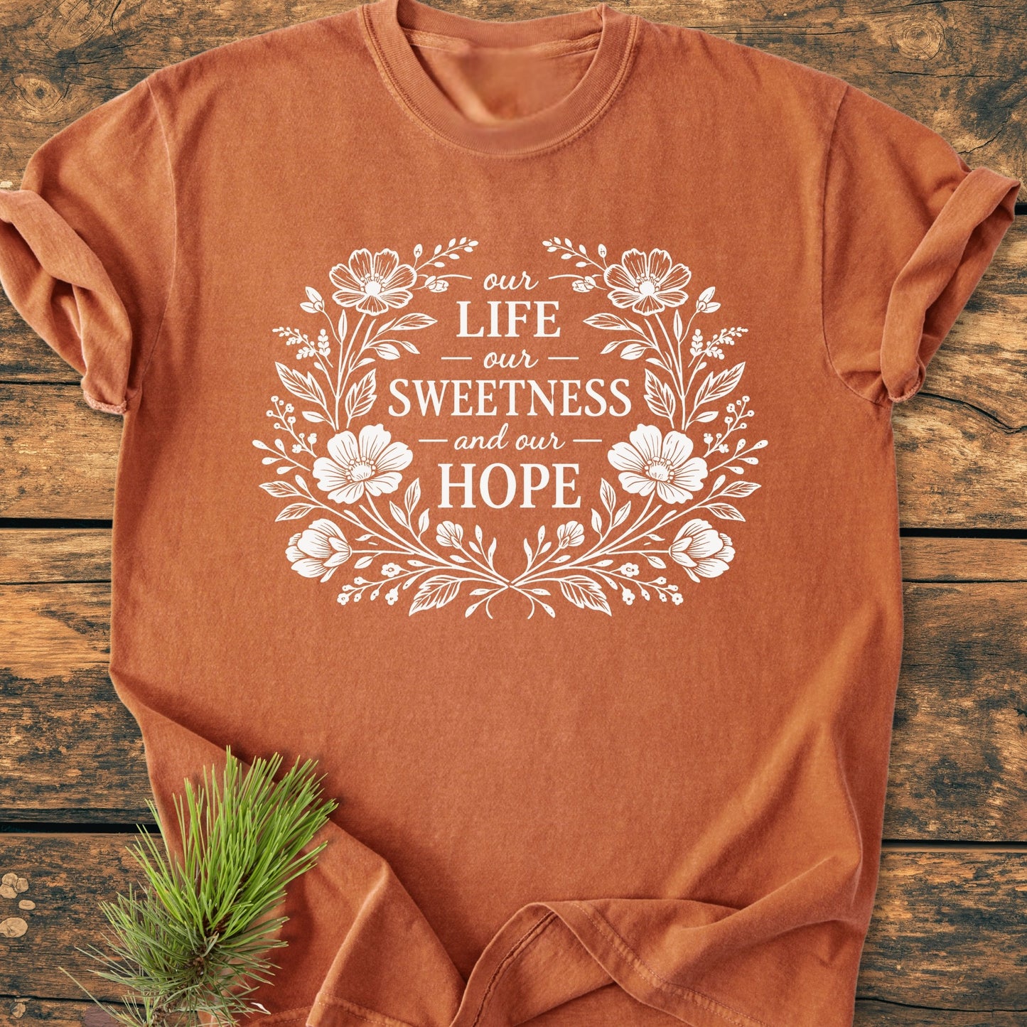 Our Life, Sweetness, Hope - Vintage Wash Tee features a floral wreath design with text, laid on wood. Pine sprig accents, rolled sleeves highlight the Comfort Colors' relaxed fit and durable stitching.
