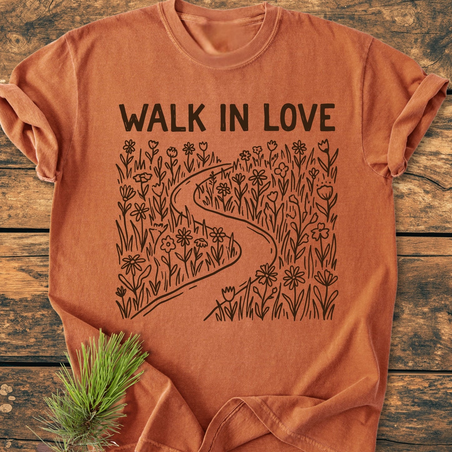 Walk In Love - Vintage Wash Tee, showcasing a burnt orange T-shirt with a black line-art of a winding path and “WALK IN LOVE” text, laid on a rustic wood surface with a pine sprig beside.
