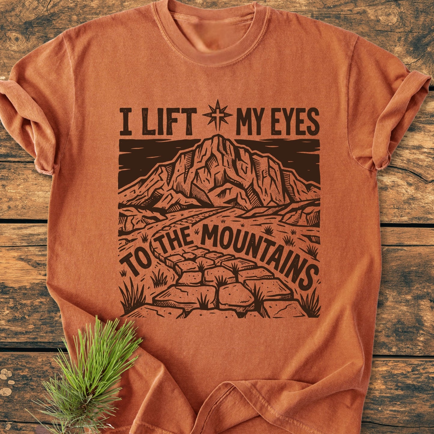 I Lift My Eyes, Psalm 121 - Vintage Wash Tee features a mountain scene and compass star on a rust-orange crew-neck, laid flat on wood with a pine sprig, highlighting its cozy, relaxed design.