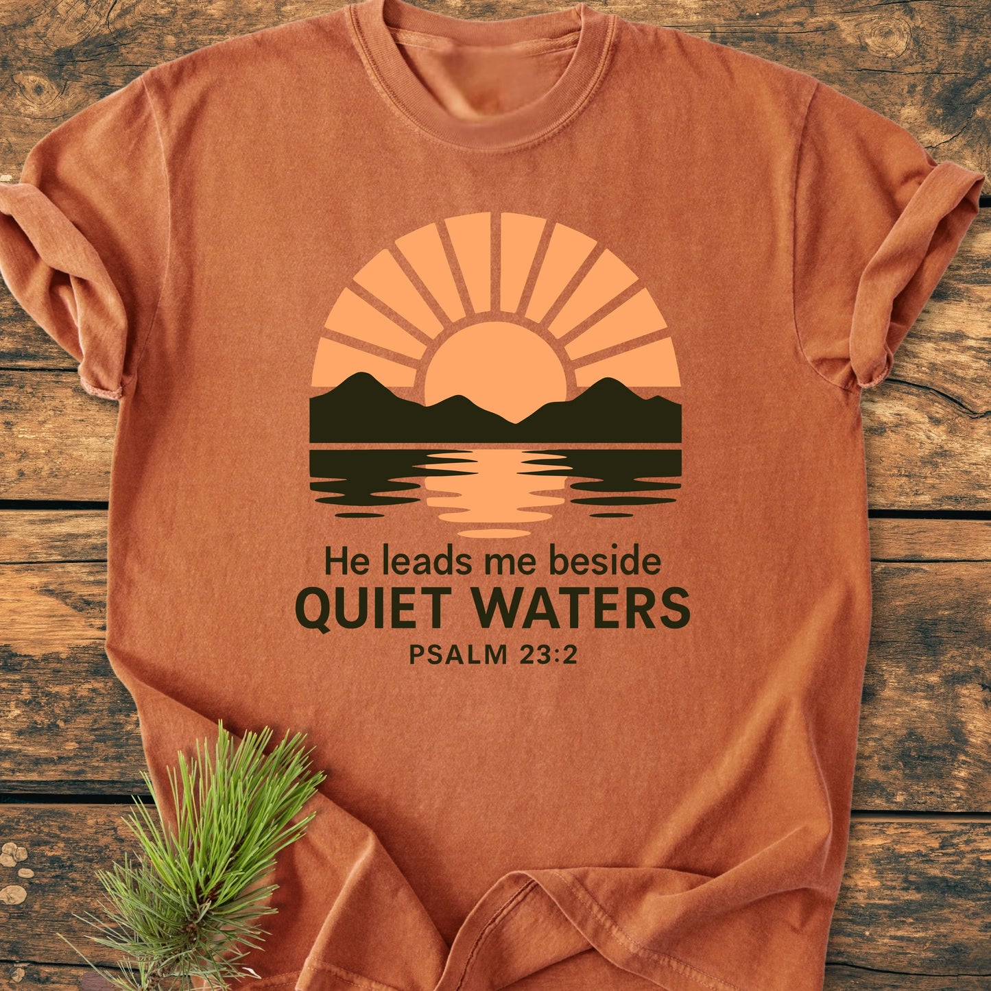 Rust-orange 1424 He Leads Me T-shirt featuring a sun rising behind green mountains, with Quiet Waters text. Laid on wood, spruce branch accents one corner, emphasizing a serene, nature-inspired design.