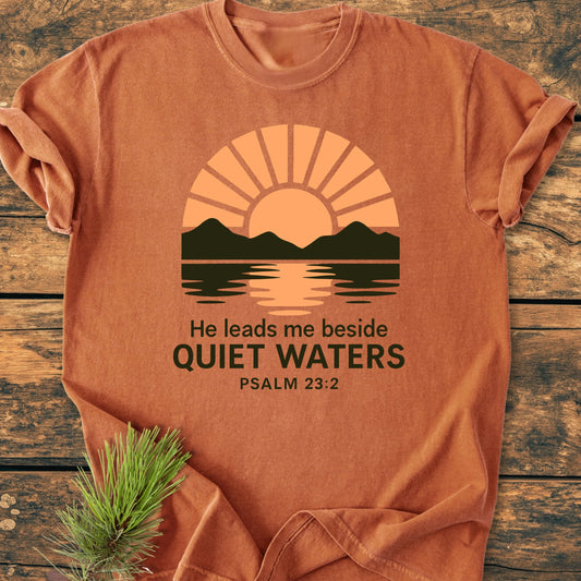 Rust-orange 1424 He Leads Me T-shirt featuring a sun rising behind green mountains, with Quiet Waters text. Laid on wood, spruce branch accents one corner, emphasizing a serene, nature-inspired design.