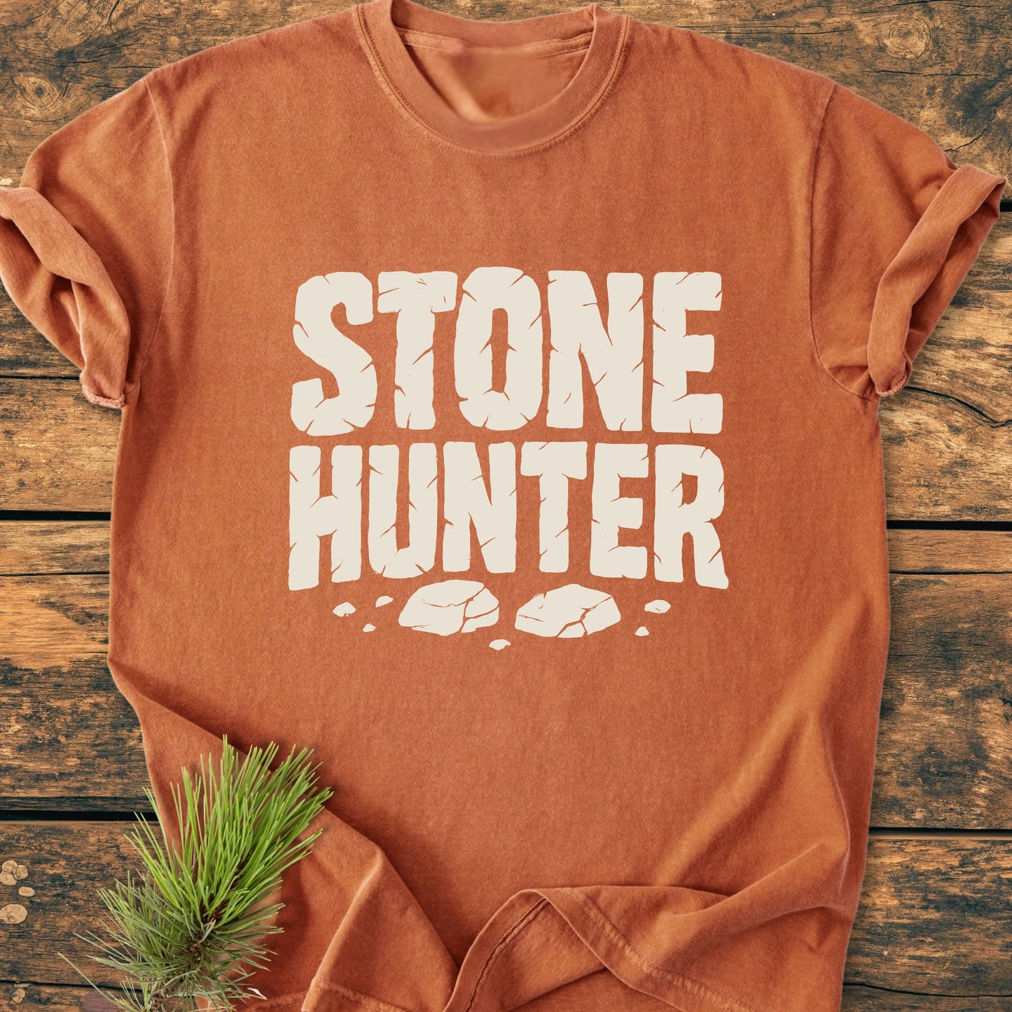 Rust-orange STONE HUNTER T-shirt with white block letters and stone graphics, laid flat on wood. Features soft vintage wash, relaxed unisex fit, and durable construction for everyday wear.