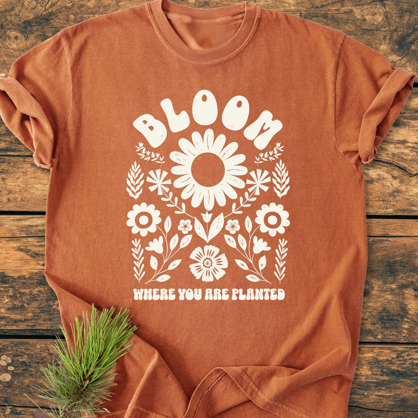 The 1415 Bloom Where You Are Planted T-shirt features a white folk-style floral graphic with daisies and foliage, and empowering text, laid on a rustic wooden surface.