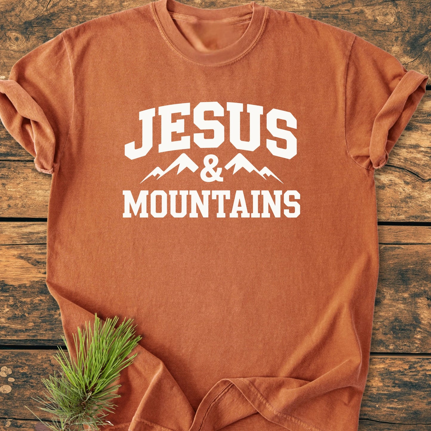 Rust-orange T-shirt with JESUS & MOUNTAINS in white, featuring a mountain graphic, laid flat on a wood surface with a pine sprig nearby, showcasing its vintage-wash, relaxed unisex design.