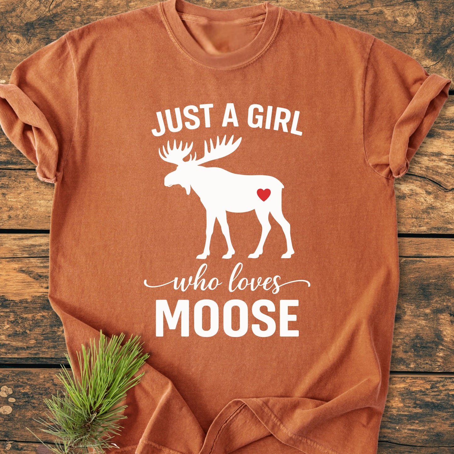 1437 Just A Girl Who Loves Moose T-shirt featuring a moose silhouette with a heart, laid on wood with a pine sprig, showcasing its vintage wash, relaxed fit, and soft, durable fabric.