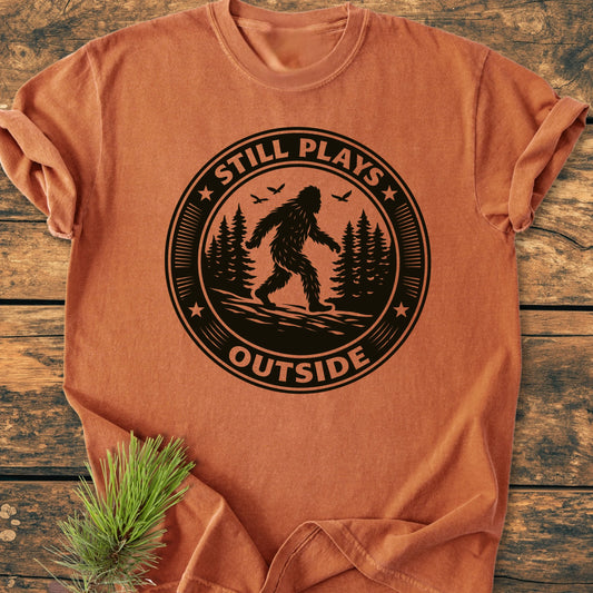 1430 Still Plays Outside Bigfoot T-shirt displays a central black emblem with Bigfoot, pine trees, and birds, encapsulated by the phrase STILL PLAYS OUTSIDE, on a relaxed, vintage-wash tee.