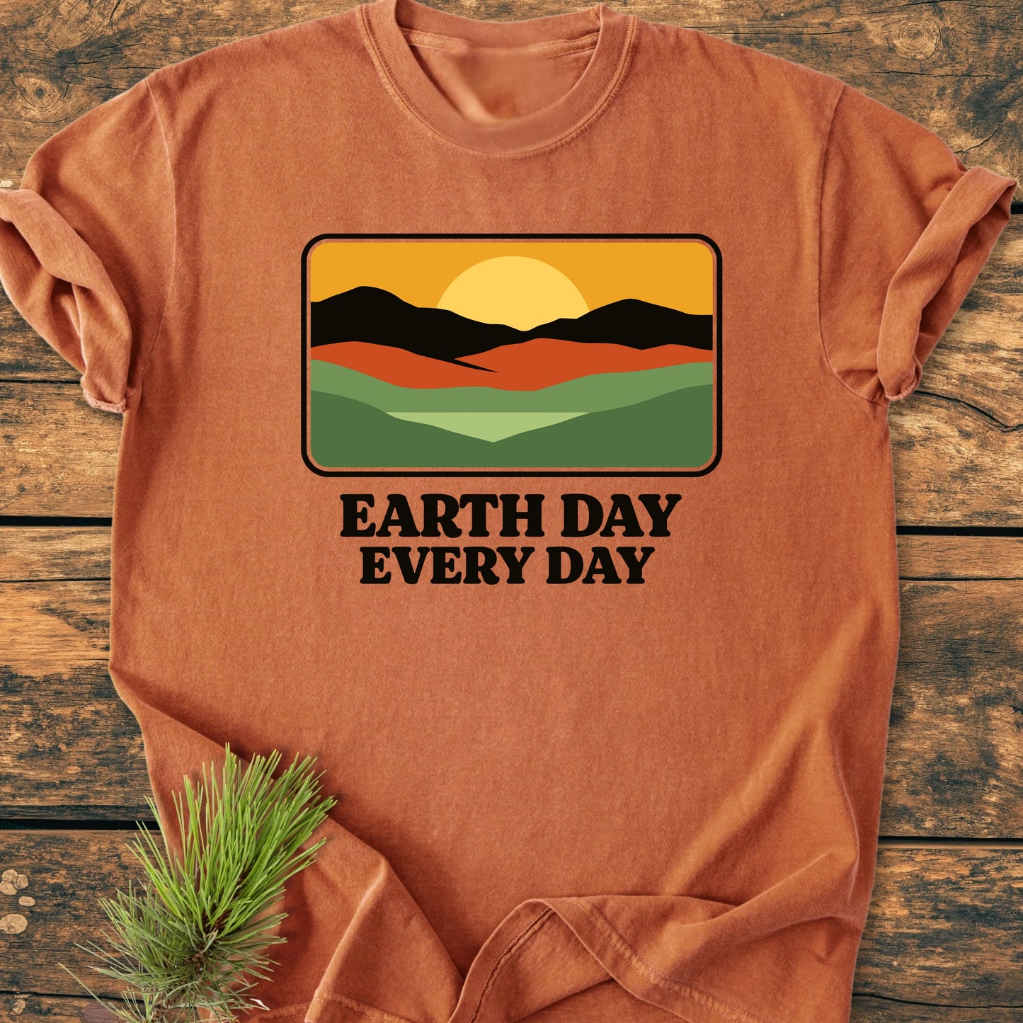 Earth Day T-shirt featuring a graphic of rolling hills with a golden sun, laid on wood. The text reads “EARTH DAY EVERY DAY.” Accompanied by a pine sprig, emphasizing the nature theme.