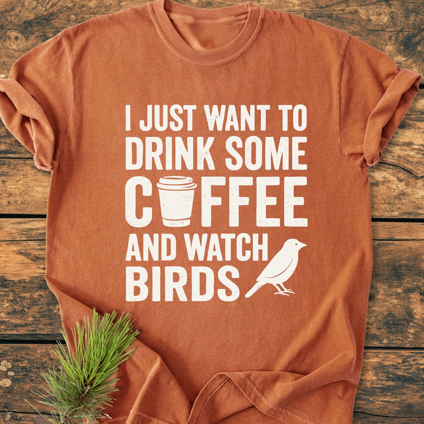 Rust-orange short-sleeved T-shirt titled 1405 Drink Coffee And Watch Birds Base, featuring bold white slogan, coffee cup icon, and bird silhouette, styled with a small pine sprig on a wooden surface.