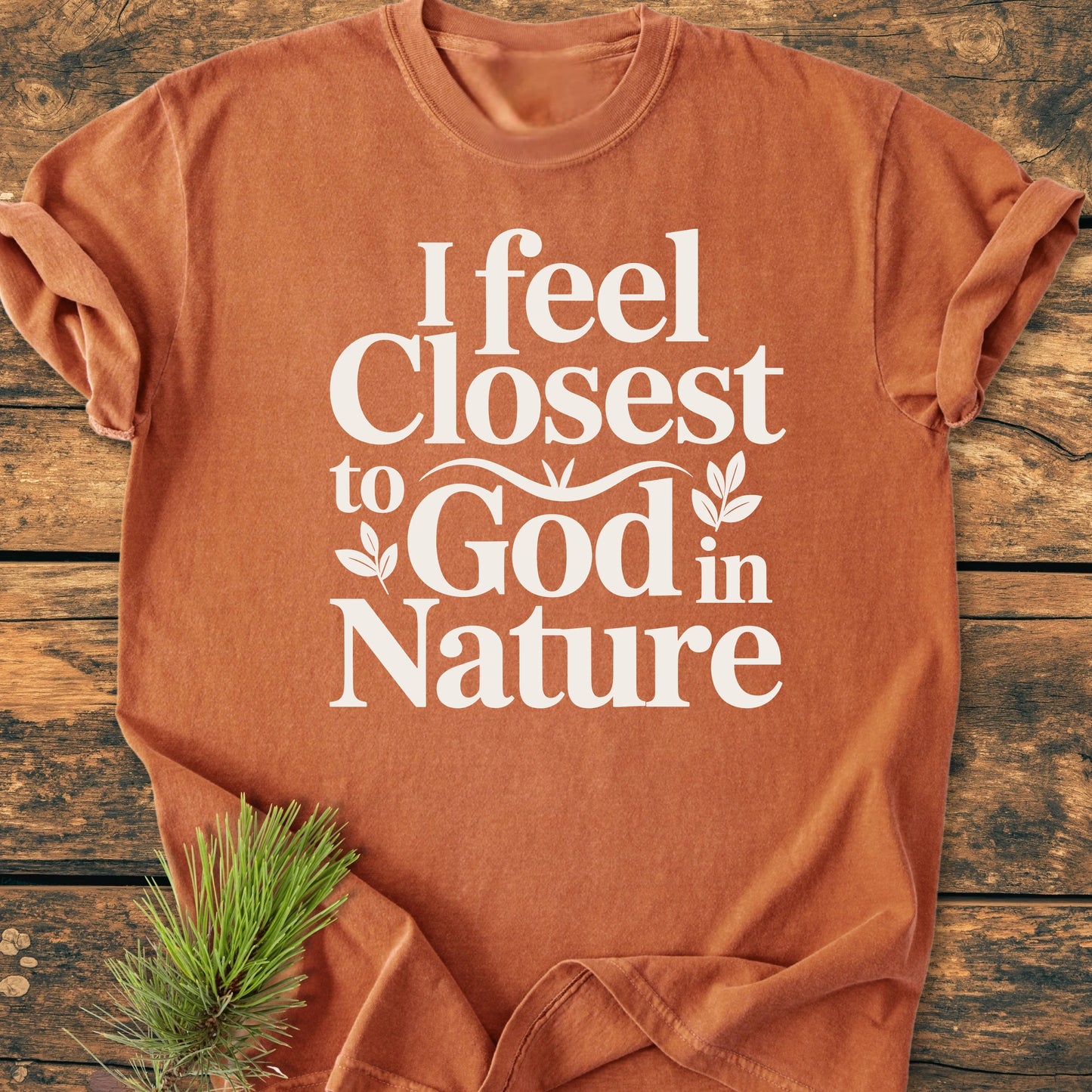 Rust-orange 1427 Closest To God - Vintage Wash Tee on wood surface, white lettering saying I feel Closest to God in Nature, with pine needles tucked under hem for an outdoorsy touch.