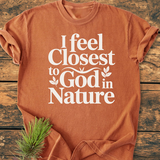 Rust-orange 1427 Closest To God - Vintage Wash Tee on wood surface, white lettering saying I feel Closest to God in Nature, with pine needles tucked under hem for an outdoorsy touch.