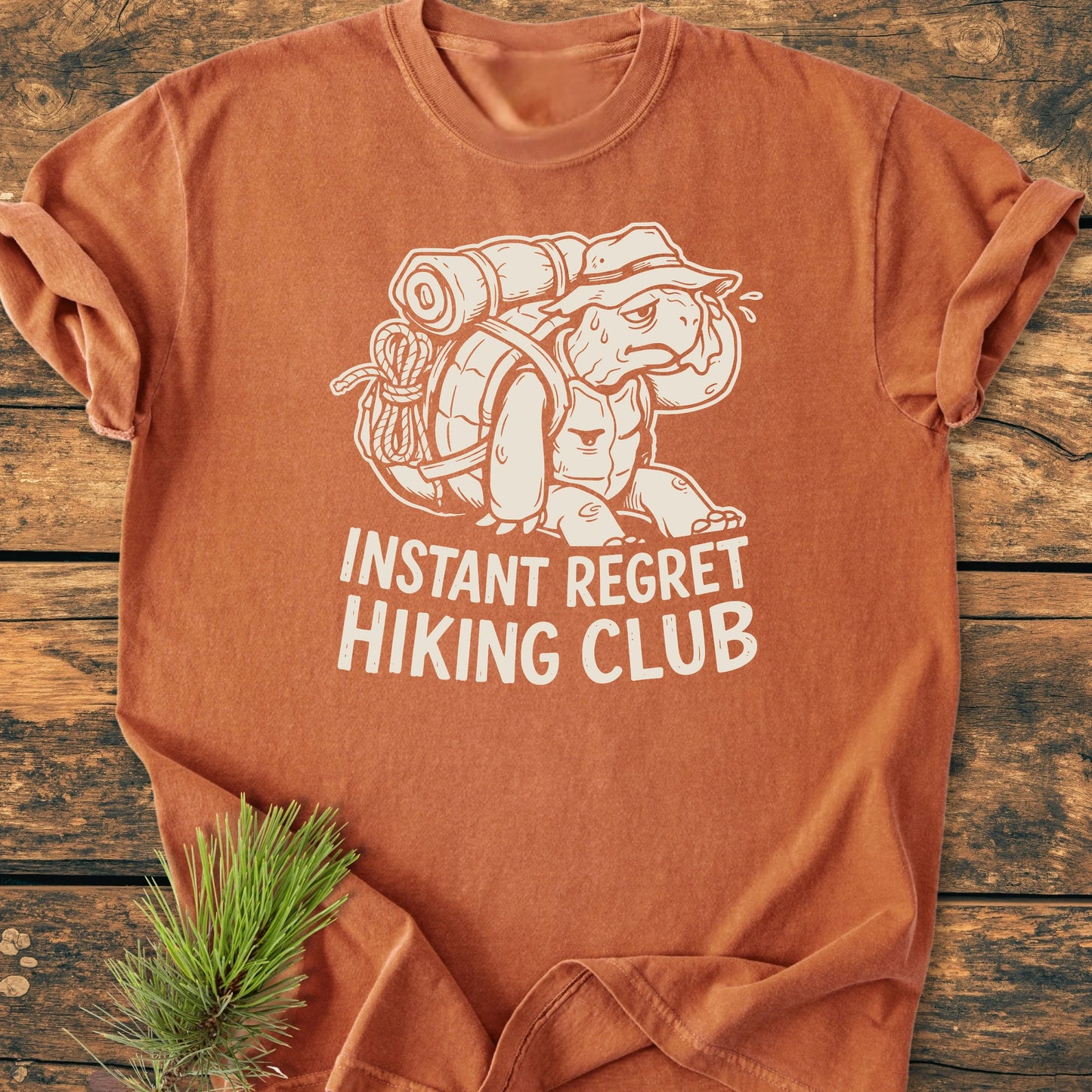 Rust-orange T-shirt featuring 1430 Instant Regret Hiking Club with a cartoon turtle in hiking gear. Laid on a wooden surface, it embodies a vintage, outdoorsy feel.