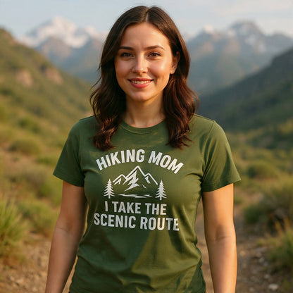 Young woman wearing the Hiking Mom, Scenic Route T-Shirt, featuring a white mountain graphic, stands smiling on a scenic dirt trail with rolling hills behind her.