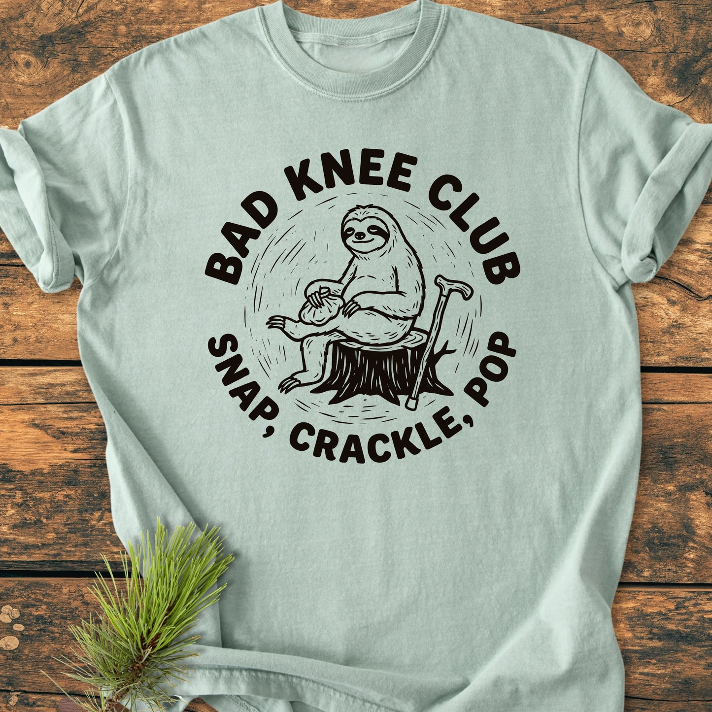 Light-green T-shirt featuring “BAD KNEE CLUB” design with a sloth on a stump, ice/heat pack on knee, and cane. Vintage wash tee, soft and relaxed fit for everyday wear.