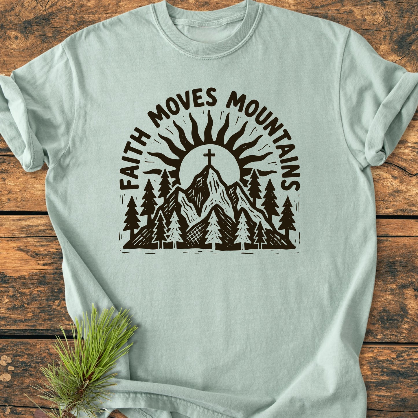 1409 Faith Moves Mountains T-shirt features a hand-drawn mountain graphic with a cross and sun, emphasizing 'FAITH MOVES MOUNTAINS' text, laid on wooden surface with pine sprig.