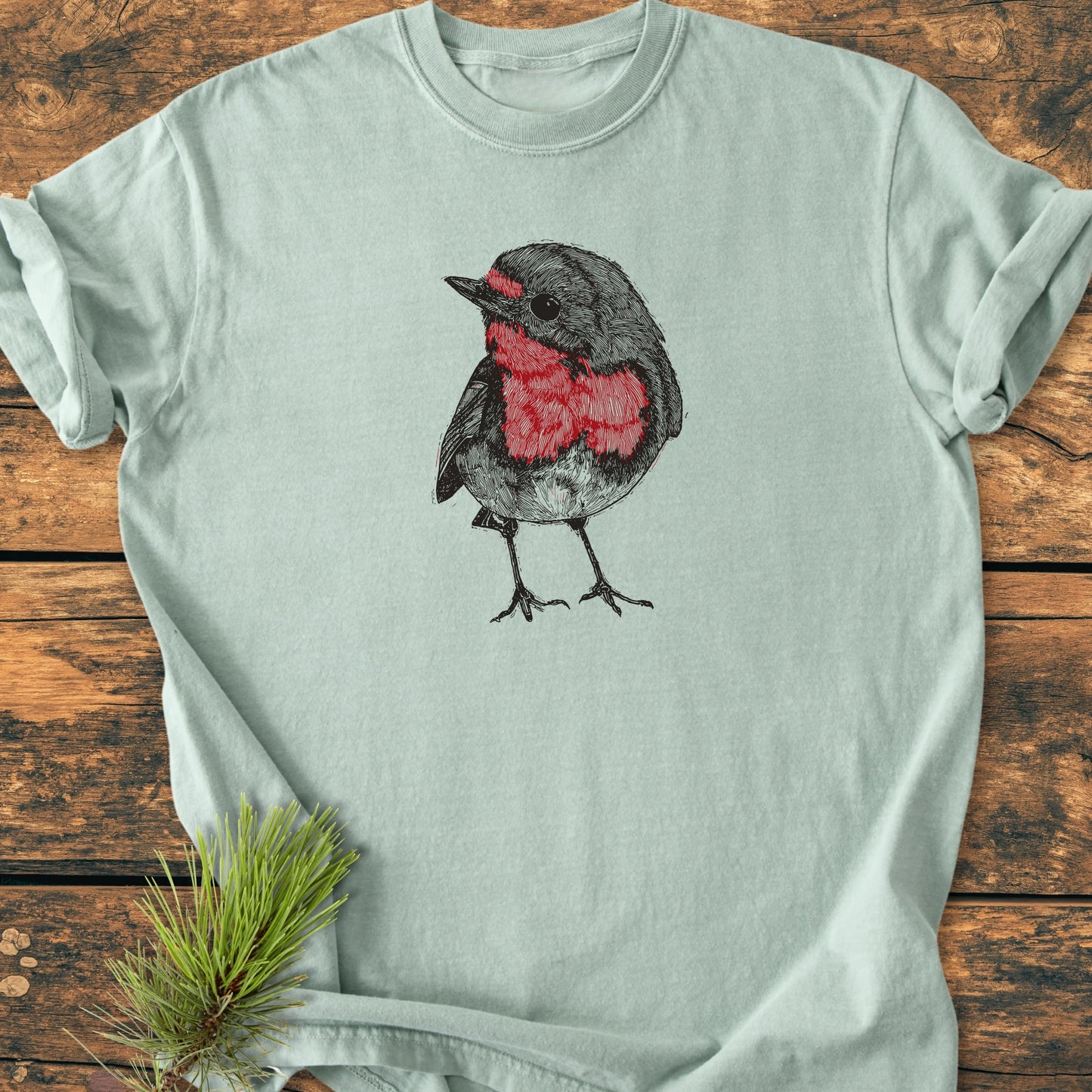 Cute Robin: A mint-green T-shirt with a hand-drawn robin illustration on the chest, displayed flat. A sprig of pine needles rests beside, enhancing the garment's natural, vintage appeal.