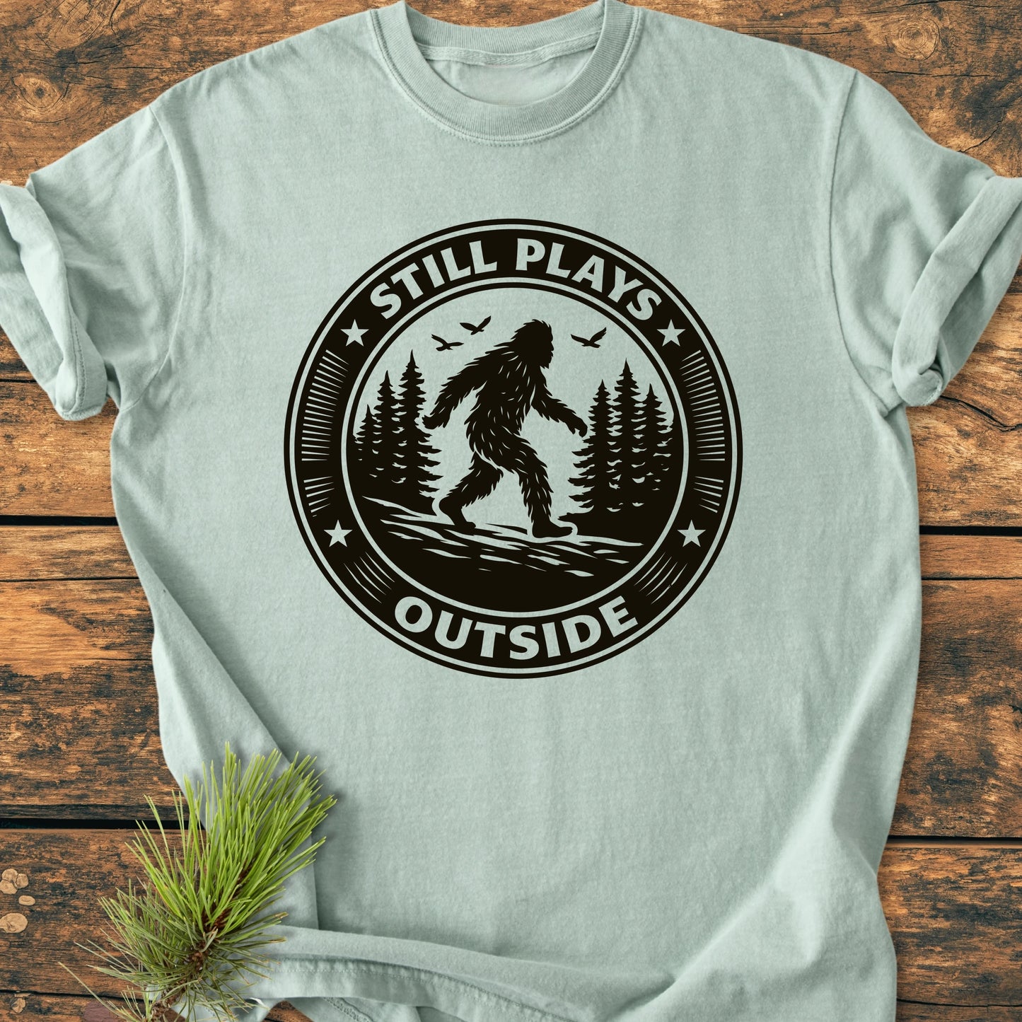 1430 Still Plays Outside Bigfoot T-shirt features a vintage-style Bigfoot graphic amidst trees, with the words “STILL PLAYS” above and “OUTSIDE” below, laid on a wooden surface.