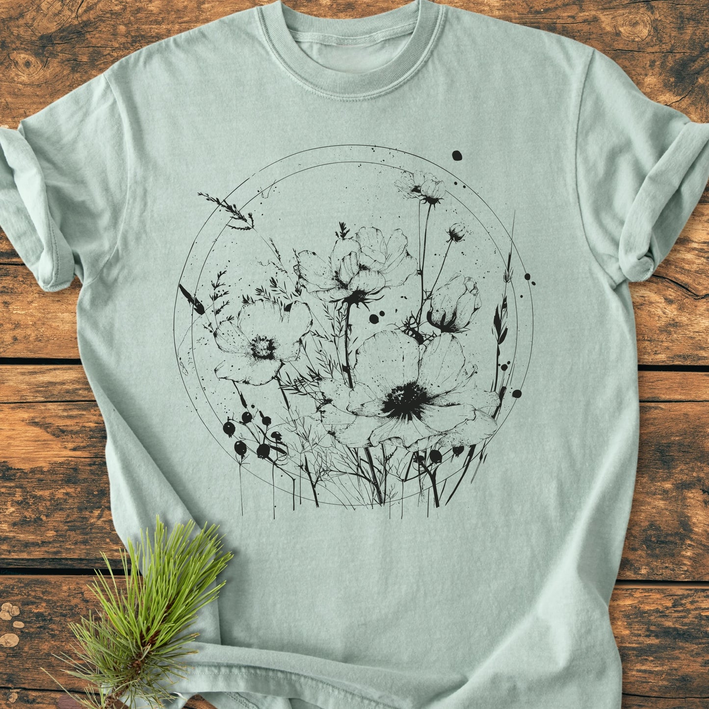 The 391 Wildflower Circle T-shirt features a black wildflower illustration within a sketched circle on a pale mint-green fabric, laid flat with a pine branch beside it.